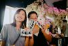 two smiling women standing in front of white flowers decorations, holding some printed photos of the night