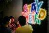 three people smiling and taking picture in front of a light sign Stay Weird